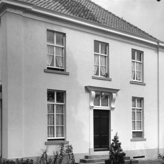 Witgepleisterd huis onder schilddak, eenvoudige, strakke lijstgevels met hoeklisenen en ramen uit de bouwtijd. Empire-deurpartij met stoep. Gepleisterde aanbouw onder zadeldak aan de achterzijde