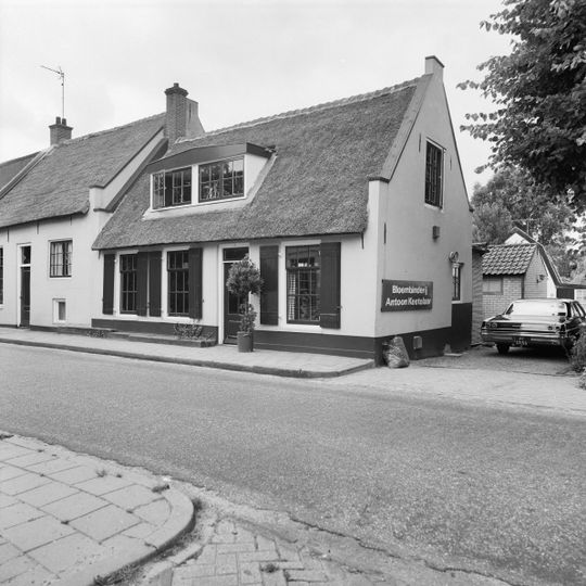 Gepleisterd dwarshuis onder rieten zadeldak tussen puntgevels. In de voorgevel vensters met luiken en negenruitsschuiframen
