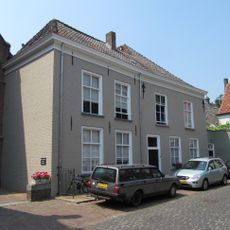 Huis op L-vormige plattegrond met grote kap, witgepleisterde lijstgevels, sierankers en gesneden bovenlicht