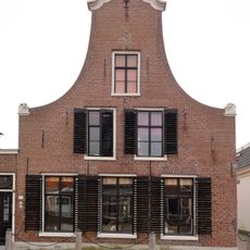 Pand met verdieping en vlieringverdieping onder zadeldak tegen slanke klokgevel met aanzetkrullen en gebeeldhouwde kuiflijst