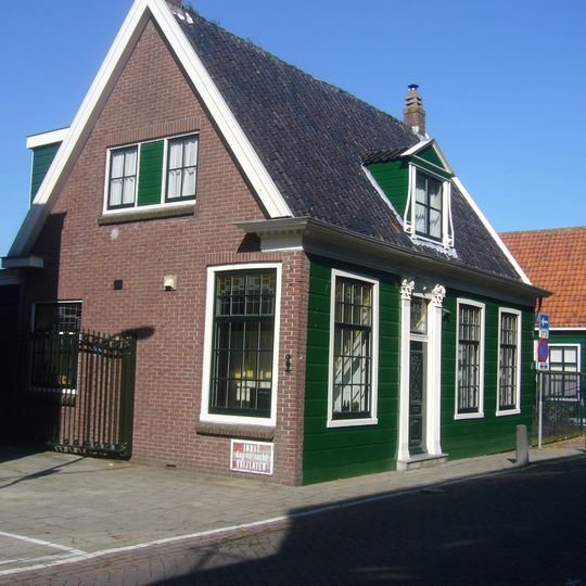 Grotendeels houten huis met zadeldak parallel aan straat. Ingang met bovenlicht tussen pilasters. Het gesneden fries tussen de pilasterkapitelen met emblemen van de koopvaardij