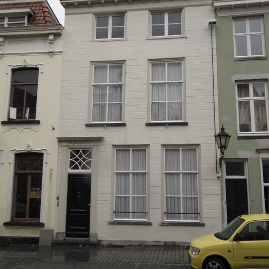 Huis met gebosseerd gepleisterde lijstgevel, kroonlijst op klossen en ingang tussen gecanneleerde pilasters. Plat dak