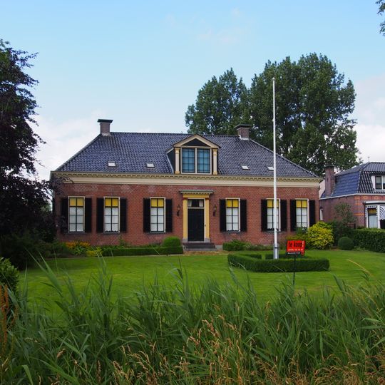 Breed landhuis met omlijste ingang, achtruitvensters en rechte kroonlijst onder laag schilddak met hoekschoorstenen met borden