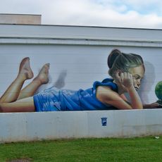 Mural de una niña vestida de azul tumbada bocabajo mientras sostiene con los ojos cerrados una maceta con una pequeña planta en forma de globo terráqueo.