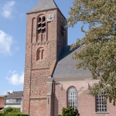Toren. Bakstenen bouwwerk, versierd tegen de onderste geledingen met tand- en boogfriezen in romaniserende trant