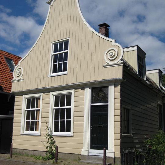 Houten dijkhuis met klokgevel uit 18de eeuw. Onderpui in empirestijl, puntgevel aan achterzijde