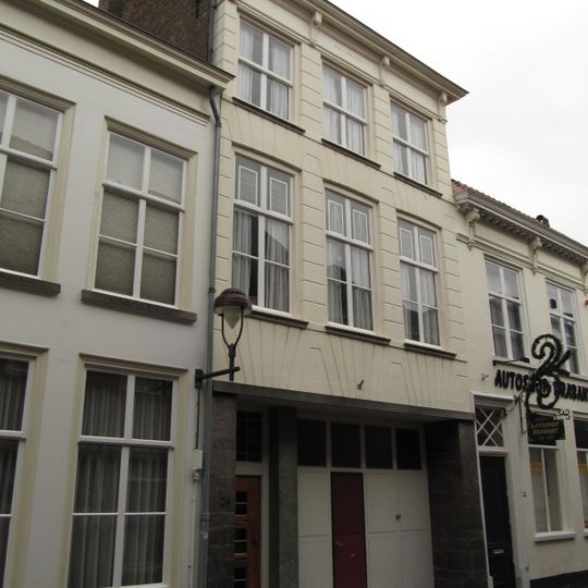 Huis met gebosseerde witgepleisterde lijstgevel, kroonlijst op voluutvormige consoles, ingang met geblokte pilasters en met kroonlijstje waarin triglyphen