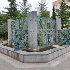 Polygonales Granit-Brunnenbecken mit vier Granitpfosten mit Petroglyphe und dazwischen gespannten Glasflächen Belmsdorfer Straße 43