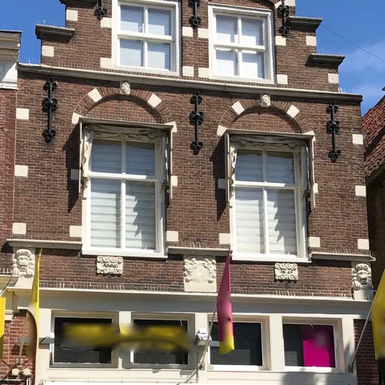 Pand onder zadeldak tegen hoge topgevel met kant-, neg- en boogblokken voor de vensternissen herbouwd in machinale steen