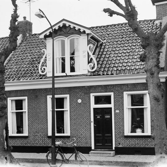 Pand zonder verdieping met voorgevel onder dwars zadeldak tussen zijtopgevels met versierde dakkapel en forse schoorsteen met bord