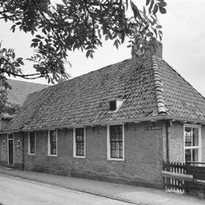 Langgerekt gebouw zonder verdieping onder zadeldak met schild aan de westzijde en aan de straatzijde een groot en vier kleine vensters met roedenverdeling