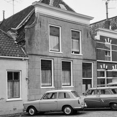 Pand onder zadeldak, aan de achterzijde afgesloten door een gepleisterde puntgevel; aan de voorzijde afgewolfd en aansluitend tegen een gepleisterde gevel met ingezwenkte zijkanten en houten kroonlijst, 19e eeuw