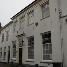 Huis met eenvoudige witgepleisterde lijstgevel met kleine roedenverdeling, in de vensters. 19e-eeuws karakter