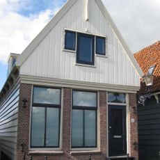 Houten huis. Voorschot in puntvorm, nog met voormalig luikkozijn met aangekoppelde vensters. Pui laatste helft 19e euew in baksteen vernieuwd