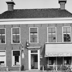 Blokvormig pand met omlijste ingang en verdieping met zesruitsvensters onder laag schilddak met twee hoekschoorstenen met borden (voorm. herberg)