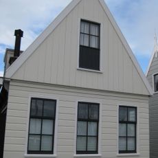 Houten huis met skelet. Puntvormig voorschot, goede pui en roedenverdeling. Gepotdekselde achtergevel met goede roedenverdeling in de vensters en een deurtje uit derde kwart 18e eeuw