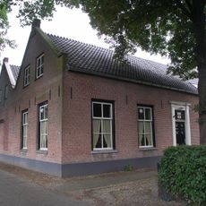 Dubbel dwarshuis, onder twee parallel lopende zadeldaken tussen puntgevels. Gesneden dubbele deur in eenvoudige omlijsting, vensters met 6-ruitsschuiframen
