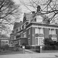 Helft van een vrijstaand bouwblok bestaande uit twee symmetrische in de diepte van een smal kavel geschakelde villa's in een historiserende bouwstijl