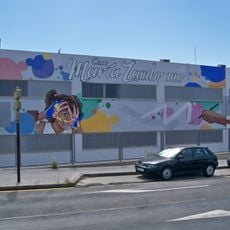 El mural muestra una mano pintando con un pincel, varias manchas de pintura de distintos colores, el texto «C. E. I. P. María Zambrano» en color blanco con bordes negro, el rostro de una niña y una mariposa monarca.